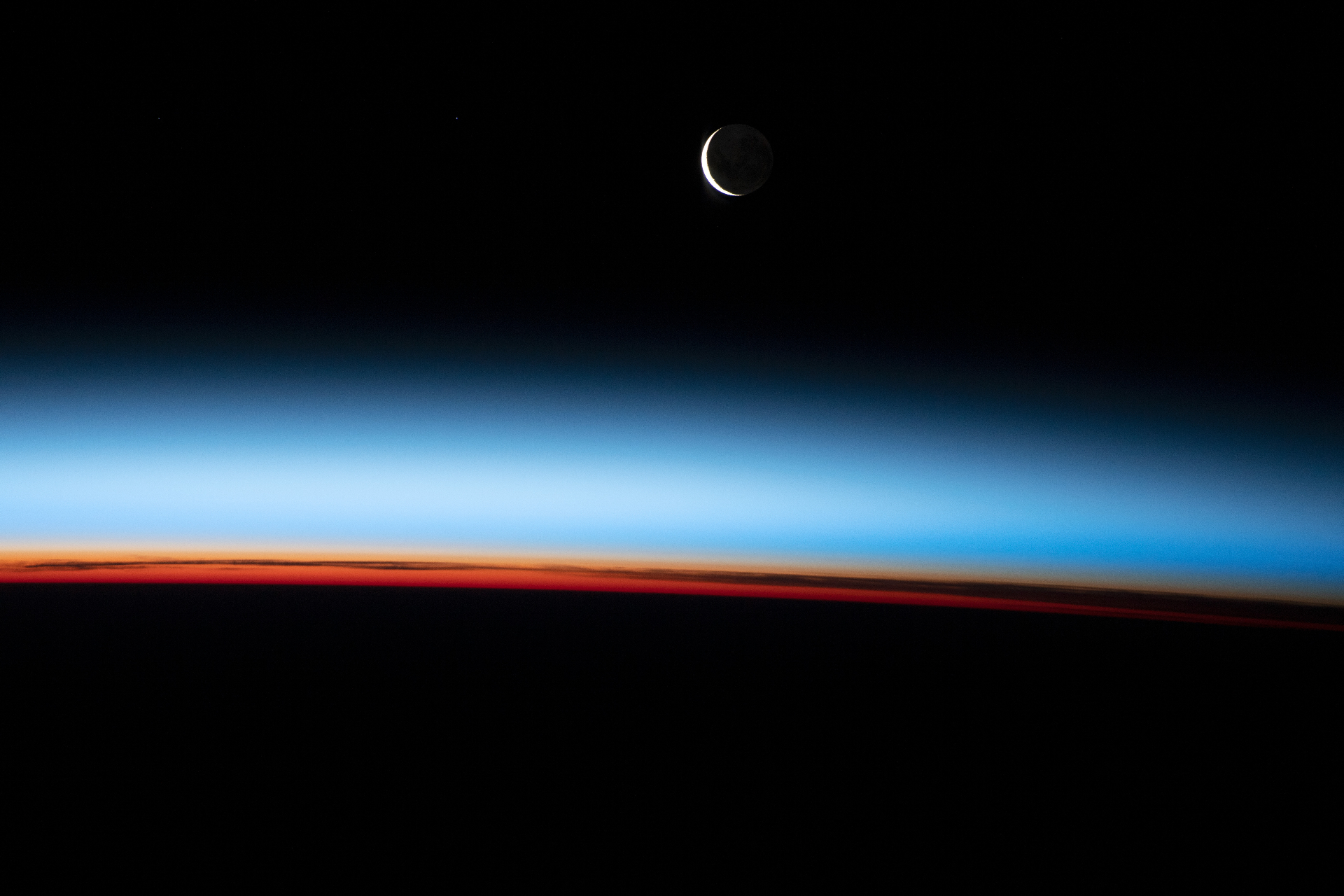 A crescent Moon above the layered colors of Earth's atmosphere at sunset, seen from orbit